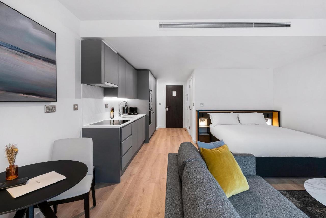 Citysuites 2 Aparthotel Manchester Exteriör bild The photo shows a modern and stylish studio apartment. It features a kitchenette with dark cabinetry and a small dining table with two chairs. There's a cozy sofa with colorful cushions, positioned next to the table. In the background, a large bed wi