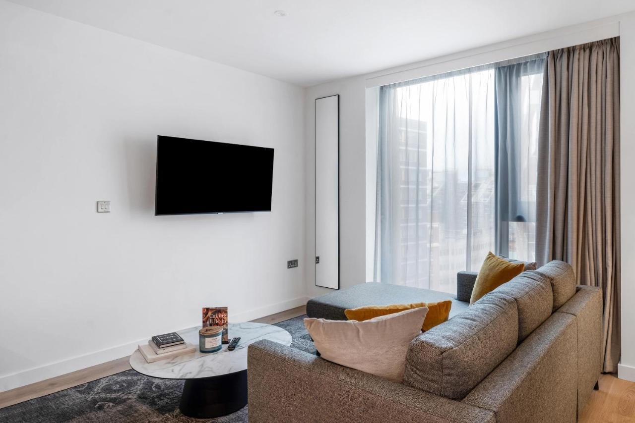 Citysuites 2 Aparthotel Manchester Exteriör bild The image shows a modern living room with a minimalist design. There is a large flat-screen TV mounted on the white wall. A comfortable gray sofa with textured cushions occupies the center of the room, complemented by a round coffee table in front of