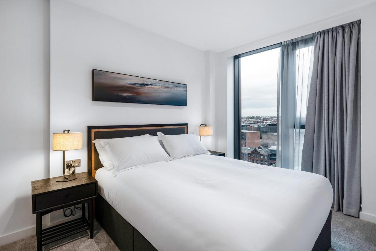 Citysuites 2 Aparthotel Manchester Exteriör bild The image shows a modern bedroom featuring a large bed with a white duvet and two pillows. On either side of the bed are small nightstands with lamps. A large window occupies one wall, draped with light gray curtains, allowing natural light to enter 