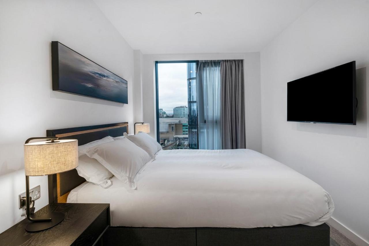 Citysuites 2 Aparthotel Manchester Exteriör bild The photo shows a modern bedroom with a minimalist aesthetic. It features a large bed with white bedding and two pillows. On either side of the bed, there are bedside tables with lamps. A large window lets in natural light, covered by sheer curtains.