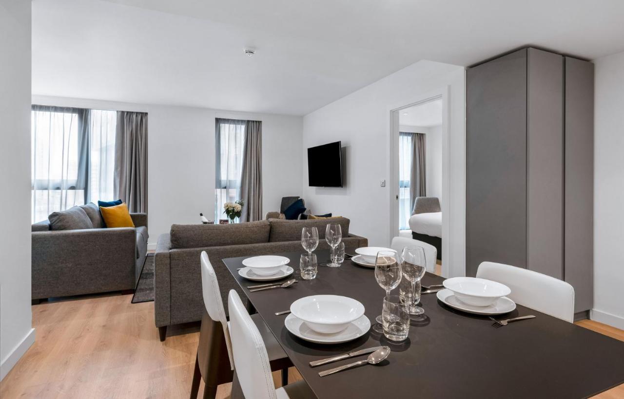 Citysuites 2 Aparthotel Manchester Exteriör bild The photo shows a modern living space with an open layout. In the foreground, there is a dining table set for a meal, featuring multiple plates, glasses, and cutlery arranged neatly. In the background, there is a comfortable-looking gray sofa with a 