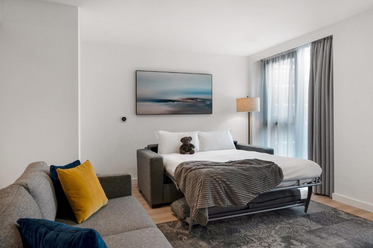 Citysuites 2 Aparthotel Manchester Exteriör bild The photo shows a modern and minimalistic guest room. There is a sofa on the left side, adorned with a colorful cushion. In the middle is a bed that appears to be a pull-out style, featuring white bedding and a gray throw blanket. A stuffed toy is pl