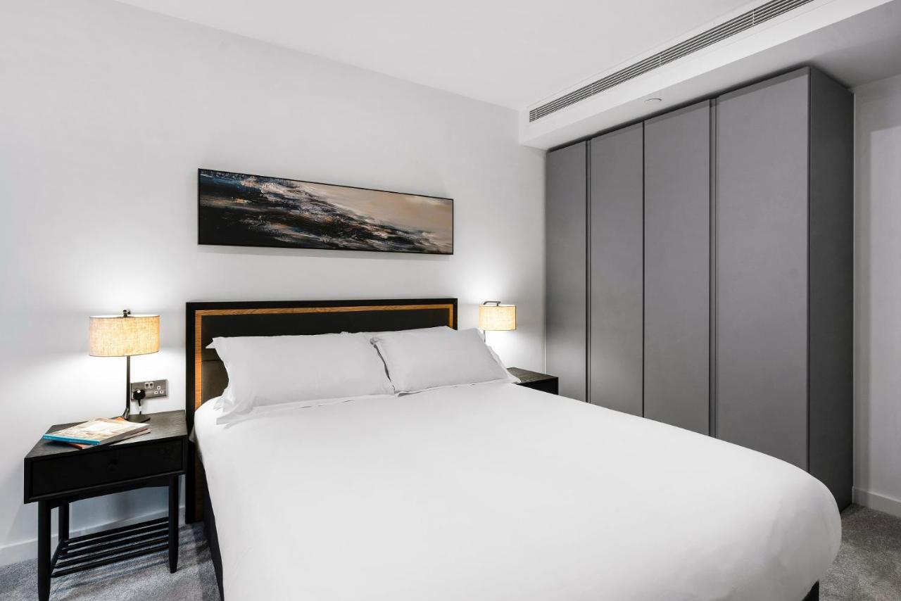 Citysuites 2 Aparthotel Manchester Exteriör bild The photo shows a modern bedroom designed with a minimalist aesthetic. The focal point is a large bed with white bedding, positioned against a wall. On either side of the bed, there are black nightstands topped with lamps, providing a warm glow. Abov