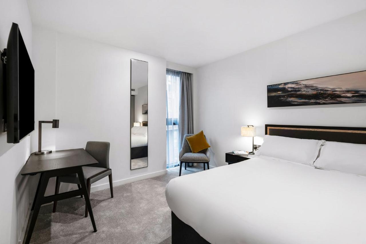 Citysuites 2 Aparthotel Manchester Exteriör bild The photo shows a modern hotel room with a clean and minimalist design. It features a large bed with white linens, a small desk with a chair, and a mirror mounted on the wall. There is a chair in one corner with a decorative pillow, and the room has 