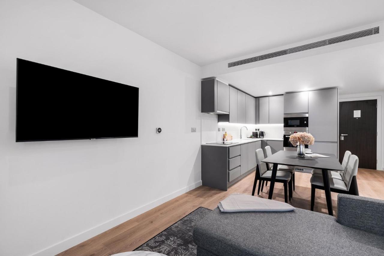 Citysuites 2 Aparthotel Manchester Exteriör bild The photo shows a modern, minimalist living space. On the left, there is a large flat-screen TV mounted on the wall. To the right, a kitchen area features sleek gray cabinets and appliances, including a built-in microwave. In front of the kitchen, th