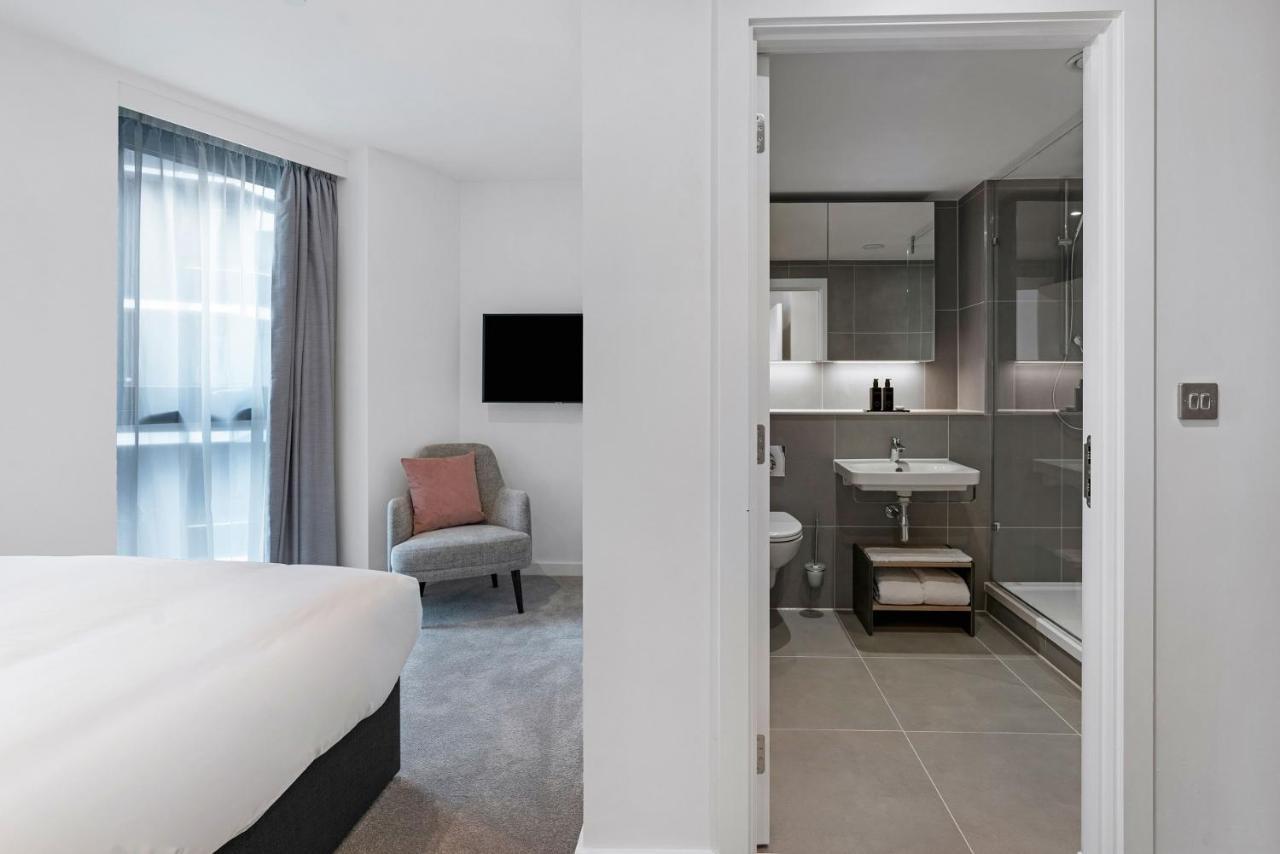 Citysuites 2 Aparthotel Manchester Exteriör bild The photo shows a modern hotel room and bathroom setup. On the left, there is a bed with white bedding and a small sitting area that includes a gray chair and a pillow in a subtle pink shade. A flat-screen TV is mounted on the wall, providing enterta