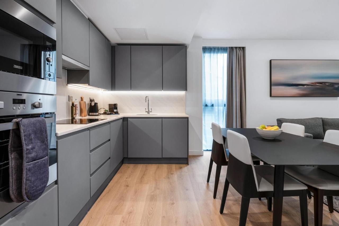 Citysuites 2 Aparthotel Manchester Exteriör bild The photo shows a modern kitchen and dining area. The kitchen features sleek gray cabinetry, a white countertop, and built-in appliances such as an oven. There is a sink and some kitchen utensils displayed. The adjacent dining area contains a black t