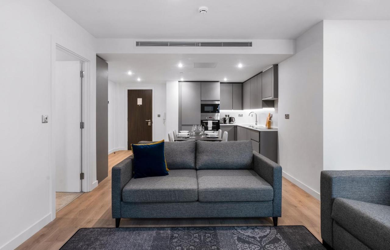 Citysuites 2 Aparthotel Manchester Exteriör bild The photo shows a modern interior space, likely a living area or apartment. In the foreground, there is a gray sofa with a blue cushion positioned on a textured rug. To the back, there is an open kitchen with sleek cabinetry and appliances, visible a