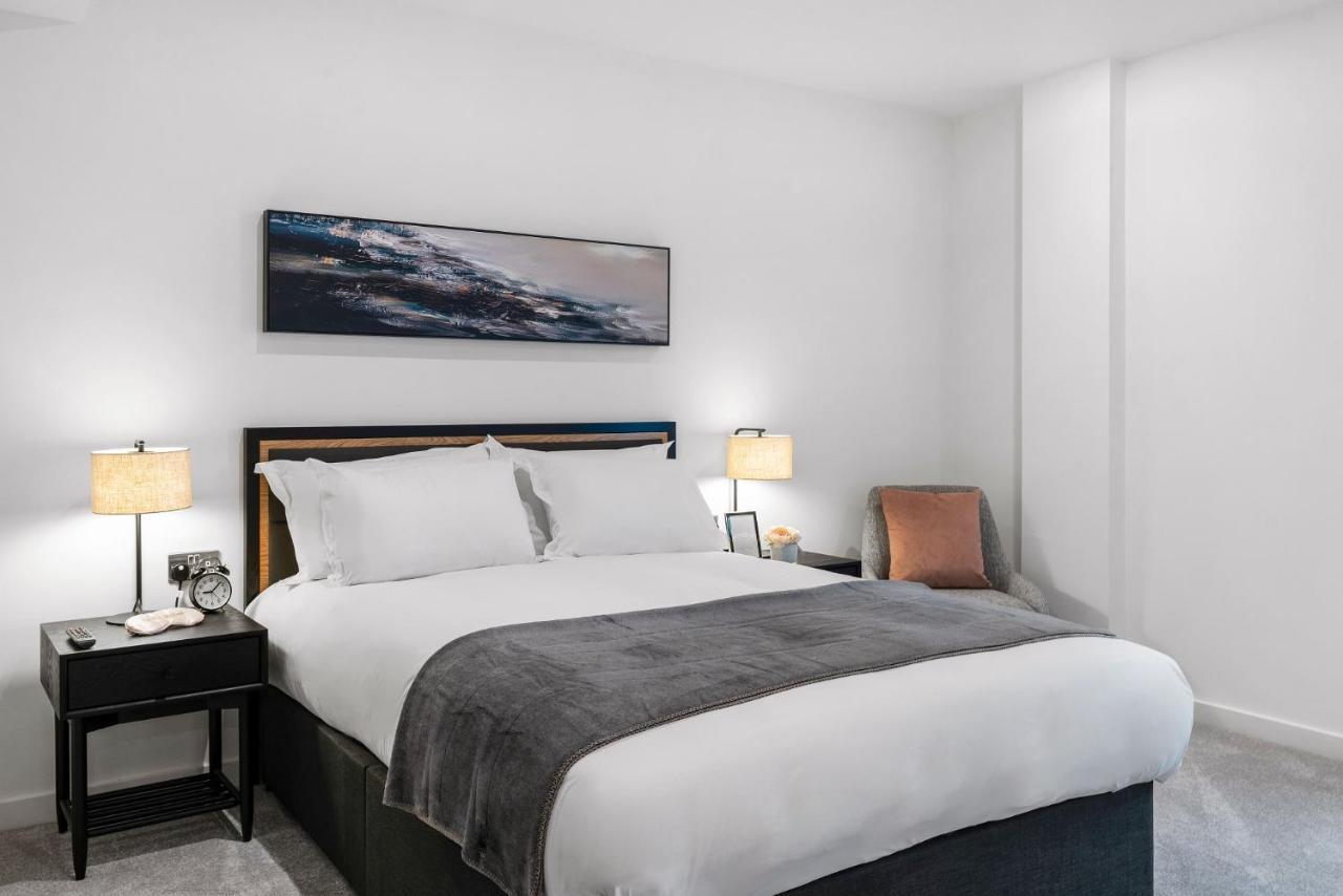 Citysuites 2 Aparthotel Manchester Exteriör bild The photo shows a modern bedroom featuring a large bed with crisp white linens and a grey throw blanket. On either side of the bed, there are bedside tables with lamps. A piece of abstract art hangs above the headboard, and there is a chair in a corn