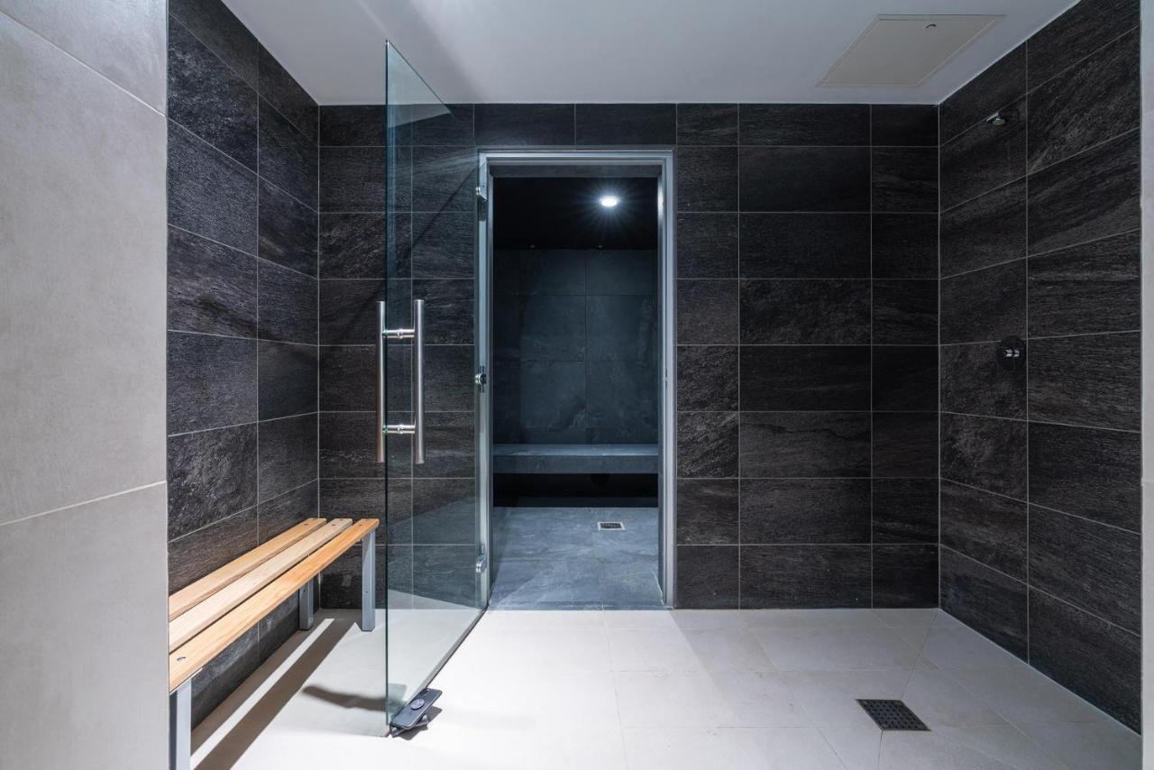 Citysuites 2 Aparthotel Manchester Exteriör bild The photo shows a modern, minimalist shower area. The space is predominantly tiled with dark gray tiles on the walls, giving it a sleek and sophisticated look. There is a glass door that leads to another room, and a wooden bench is positioned against