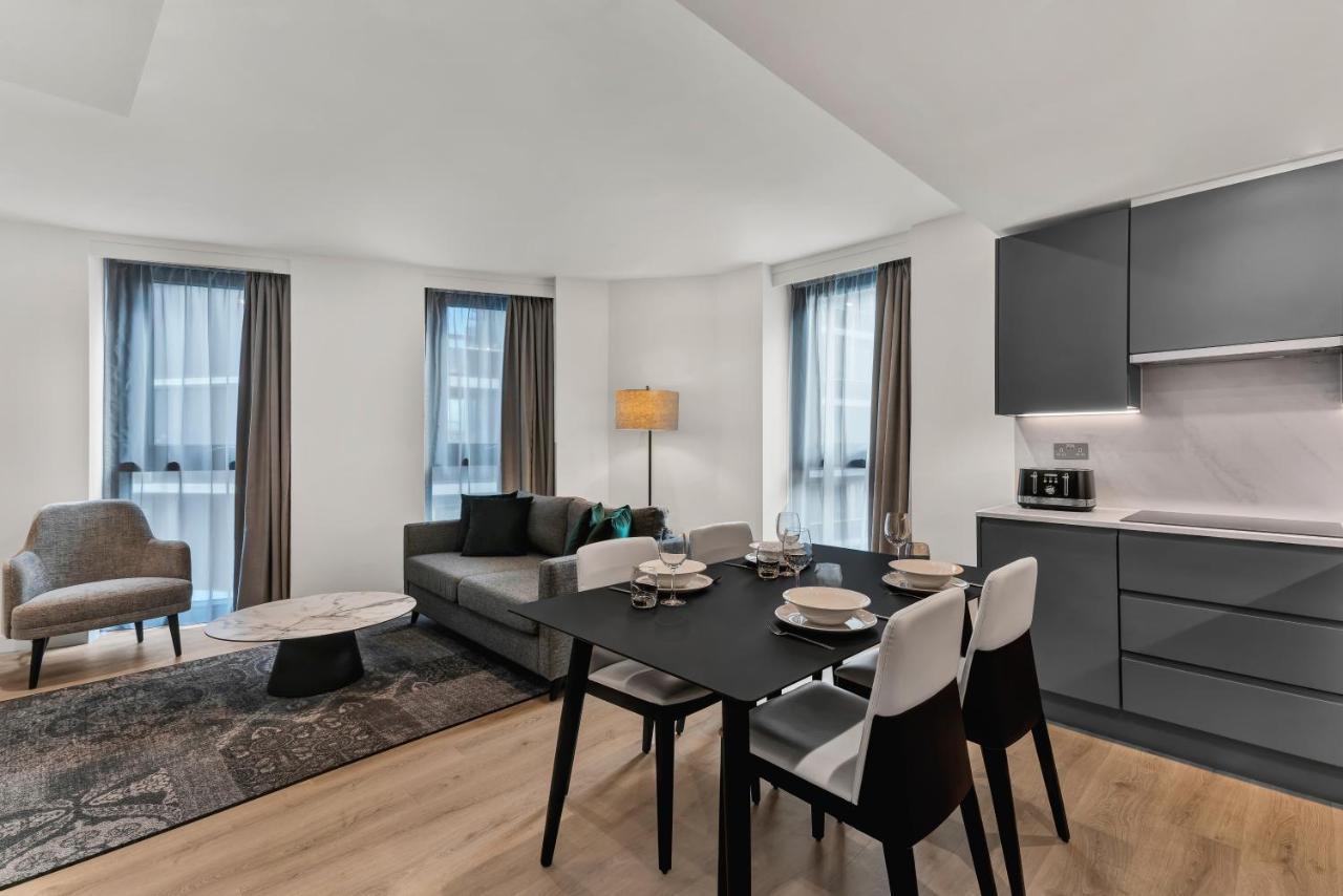 Citysuites 2 Aparthotel Manchester Exteriör bild The photo shows a modern living space that includes a dining area and a seating area. There's a sleek black dining table set with plates and cutlery, accompanied by white and black chairs. To one side, a comfortable gray sofa faces the dining area, c