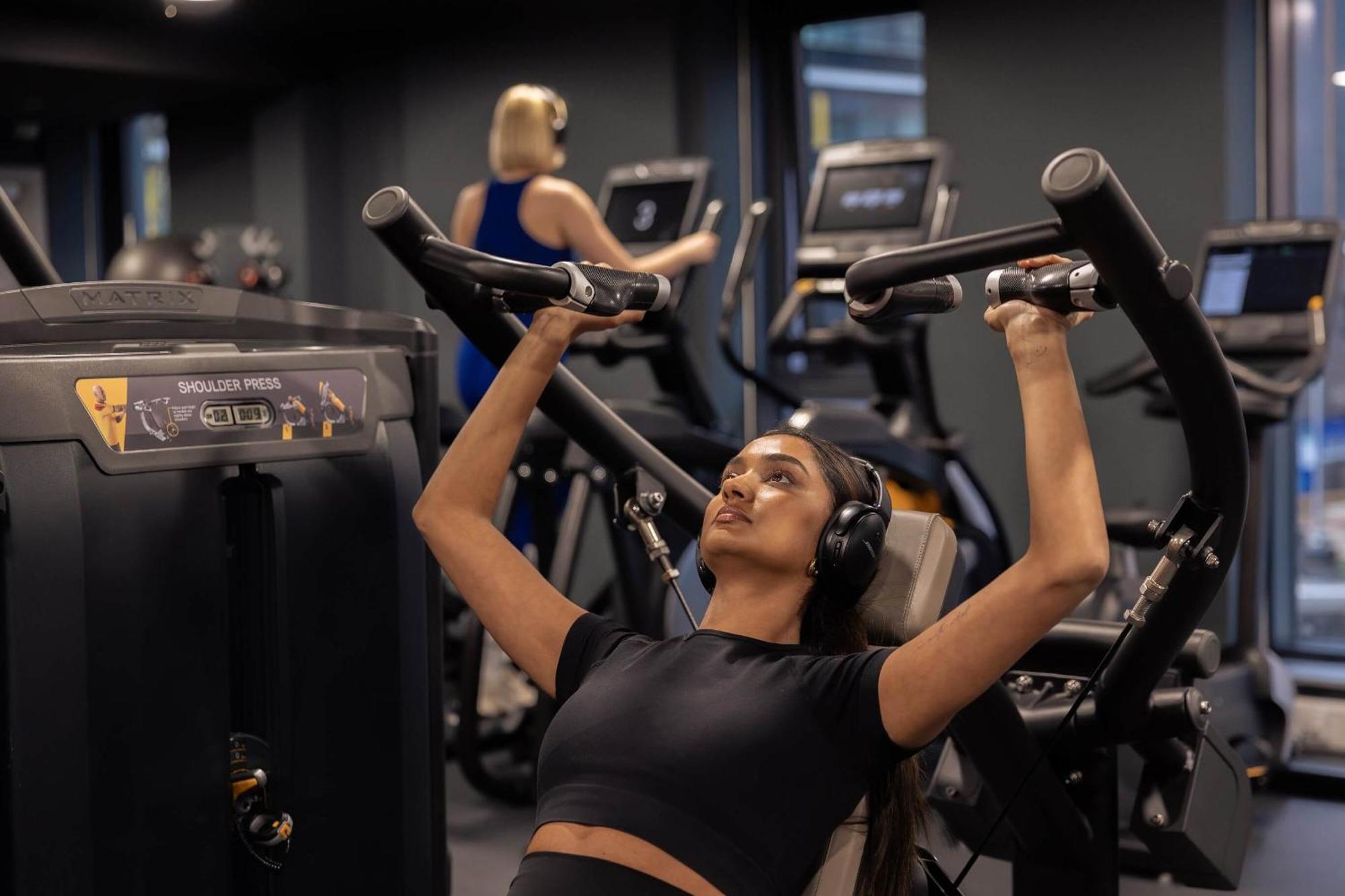 Citysuites 2 Aparthotel Manchester Exteriör bild The photo shows a gym setting with two individuals engaged in physical exercise. In the foreground, a woman is using a chest press machine while wearing headphones, focusing on her workout. She appears to be in a seated position, pushing the handles 