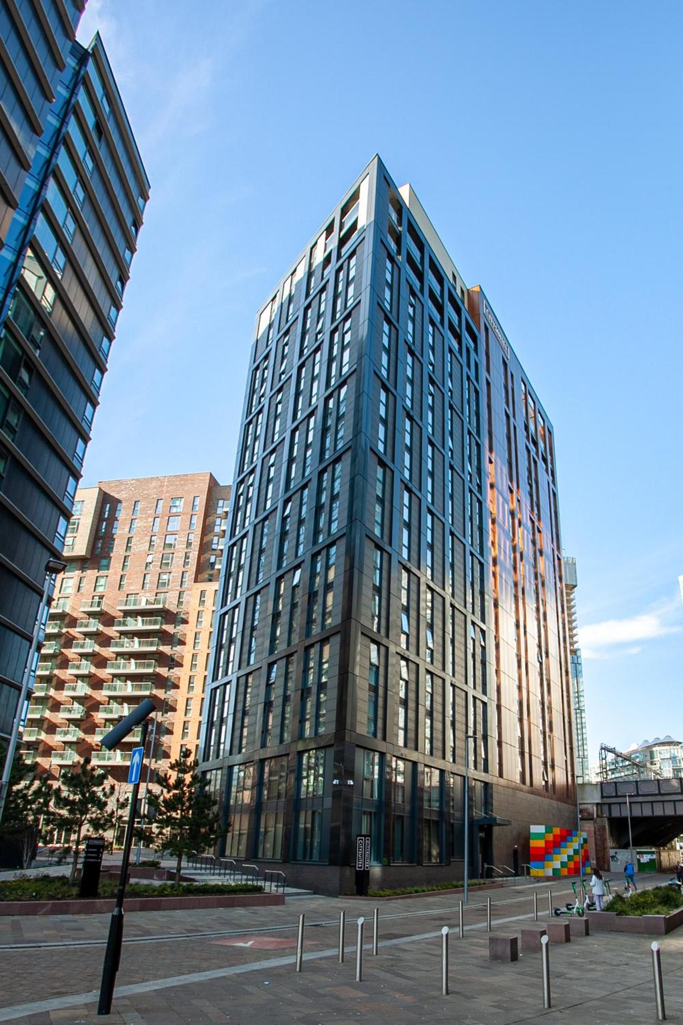 Citysuites 2 Aparthotel Manchester Exteriör bild The photo shows a modern high-rise building with a sleek, glass façade. It is set against a blue sky, indicating a clear day. The building appears to have multiple floors with large windows that reflect light. Nearby, there are additional buildings, 